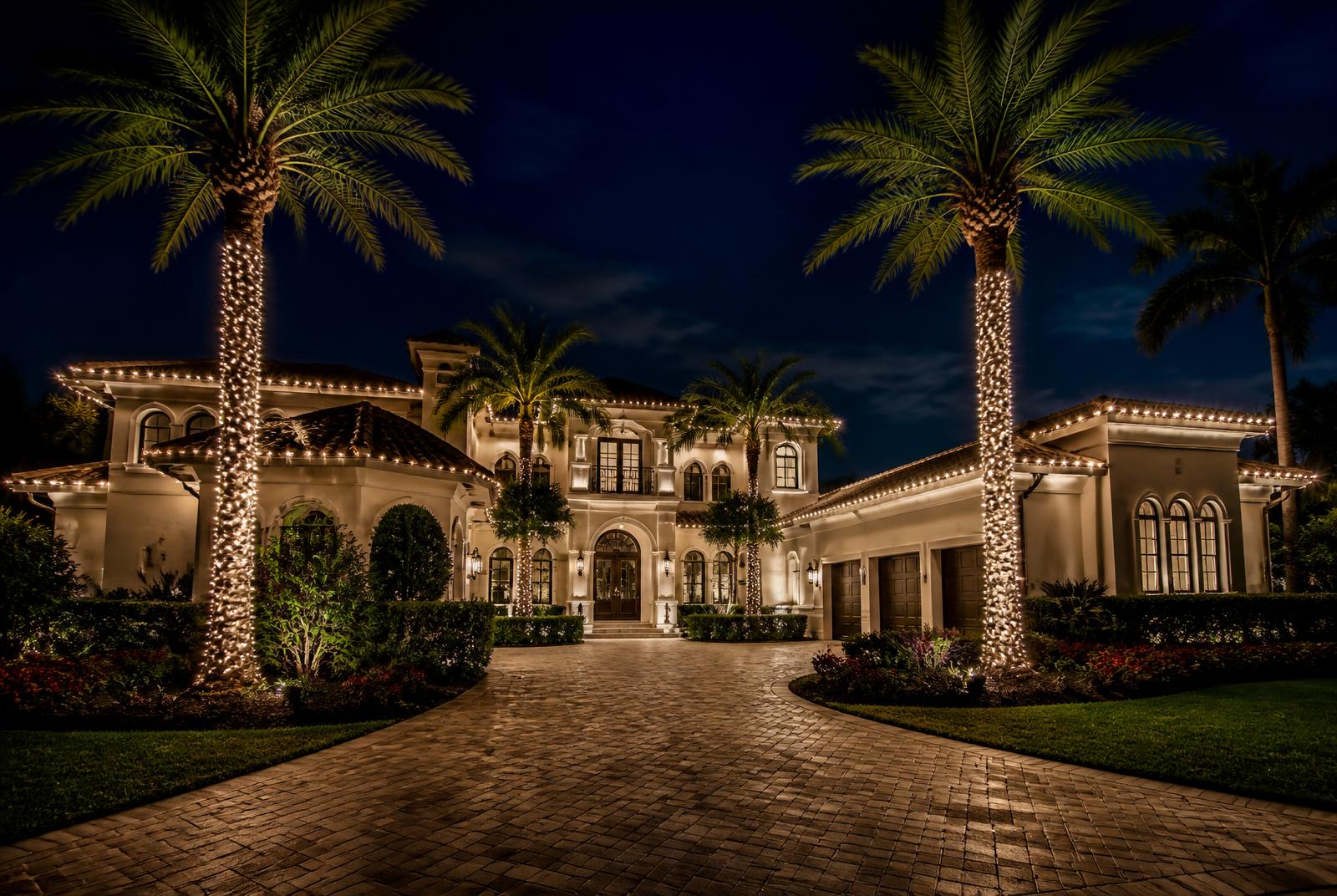Luxury Florida estate at night with warm white roofline lighting and palm trunks wrapped in lights