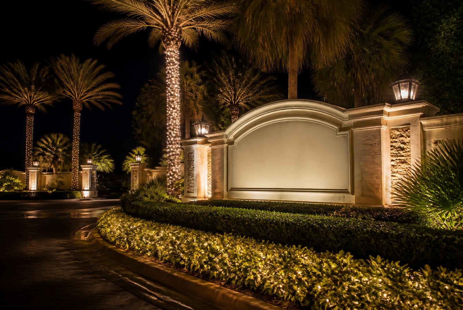 Community entrance monument at night with lit hedges and wrapped palm trunks