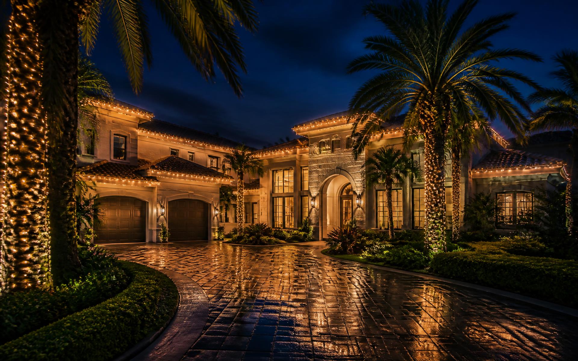 Upscale South Florida home at night with warm white lighting along the roofline and softly lit palm trees in tropical landscaping